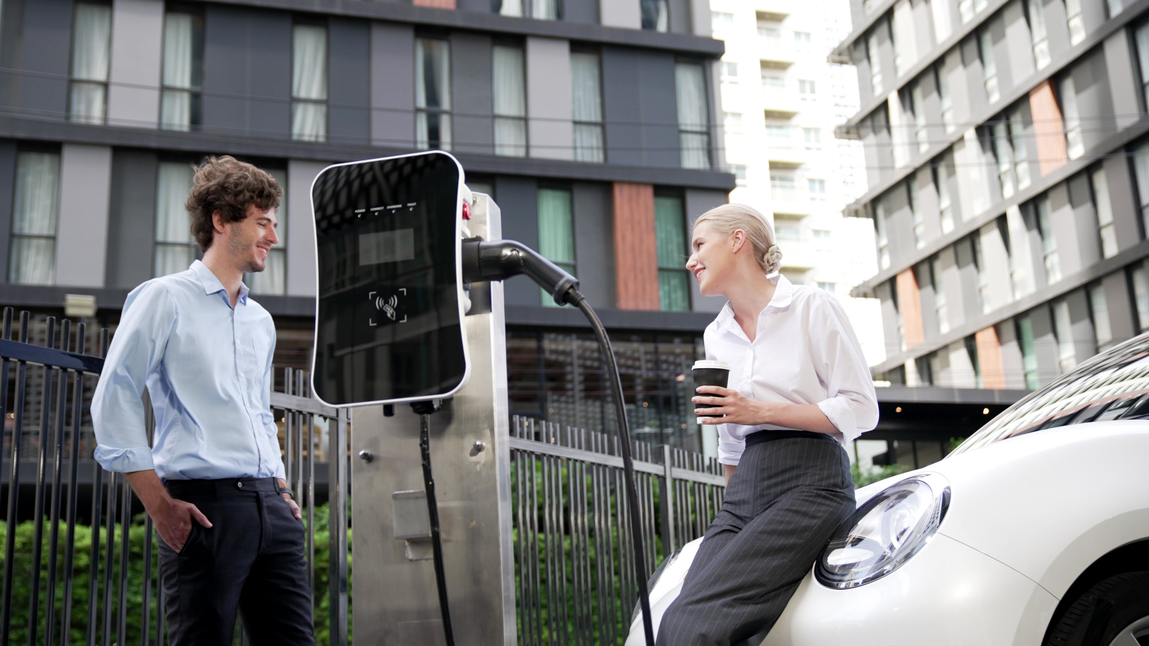 Businessman and businesswoman at an EV charging station, smiling and engaging in conversation while charging an electric car. The modern urban background features stylish buildings, emphasizing sustainable energy and electric vehicle technology